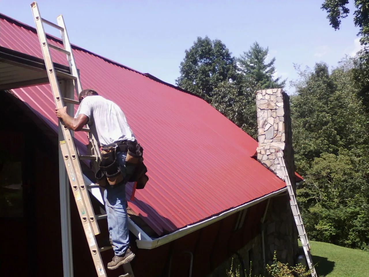 Roofer on a ladder beside a red metal roof and stone chimney — Soffit & Fascia Repair by Roofing Brothers in Owenton